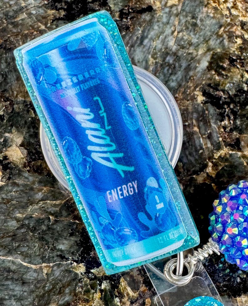 May include: A blue and teal badge reel featuring a miniature replica of an energy drink can. The can design includes the word "Alani" in white script, with "Energy" below. The badge reel has a clear plastic backing and a glittery teal border.