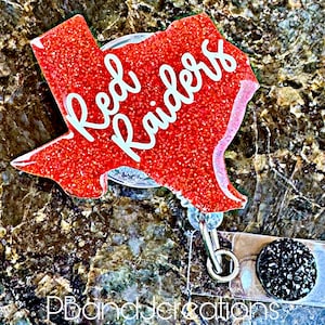 May include: A red glitter badge reel in the shape of Texas with the words "Red Raiders" in white script. The badge reel has a clear plastic base with a silver clip and a black gem-studded button.