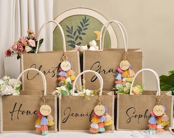 May include: Six burlap tote bags with white handles, each personalized with a name in black script. Each bag has a colorful tassel keychain. The bags are filled with flowers, suggesting a gift or party favor.