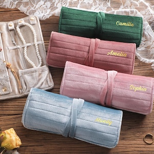 Puede incluir: Rollos de joyería de terciopelo en varios colores, incluyendo verde, rosa y azul, con nombres bordados. También se ve un organizador de joyas beige con anillos, pendientes y collares. Los rollos se cierran con una cinta de tela a juego.