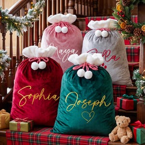 May include: Four velvet Santa sacks in red, green, pink, and gray, each featuring a white plush cuff, red and white drawstring with pom-poms, and personalized names in script font. The names Maya, Ella, Sophia, and Joseph are printed on the bags.