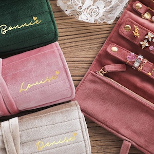 Puede incluir: Rollos de joyería personalizados en terciopelo verde, rosa y beige. Los rollos llevan nombres dorados como "Bonnie" y "Louise". Un rollo de joyería rosa abierto muestra anillos y pendientes. Un accesorio elegante y práctico.