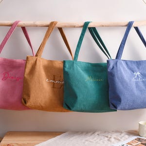 May include: Four personalized tote bags in pink, tan, teal, and blue hang from a wooden rod. Each bag has a name embroidered in a contrasting color. The bags are made of a soft fabric and have matching handles.