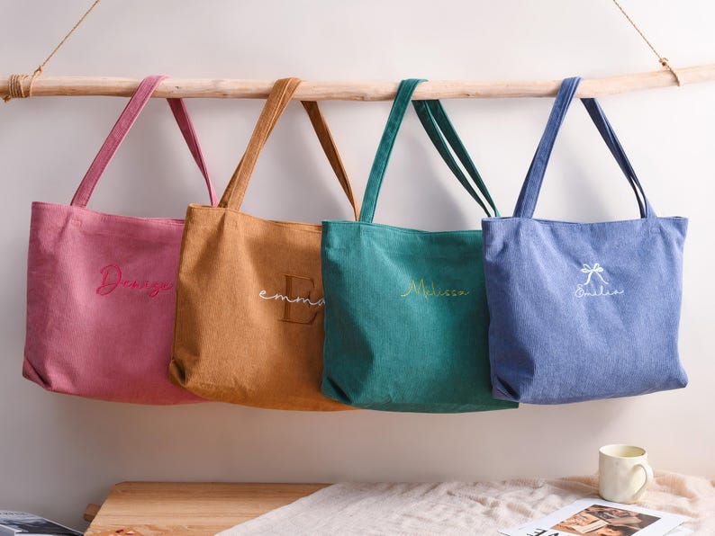 May include: Four personalized tote bags in pink, tan, teal, and blue hang from a wooden rod. Each bag has a name embroidered in a contrasting color. The bags are made of a soft fabric and have matching handles.