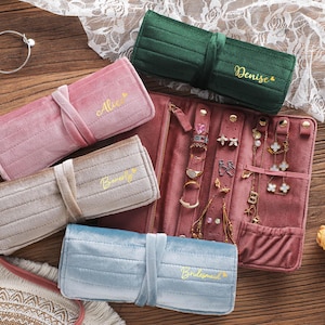 Puede incluir: Colección de organizadores de joyas de terciopelo en varios colores, incluyendo rosa, verde, beige y azul. Cada estuche enrollable está personalizado con un nombre en escritura dorada. Un estuche de joyería más grande y abierto exhibe anillos y pendientes.