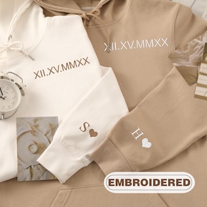 May include: Two beige hooded sweatshirts with white embroidered text. One sweatshirt has the text "XII.XV.MMXX" and the other has the text "II.XV.MMXX". The sweatshirts also have the letters "S" and "H" embroidered on the sleeves.