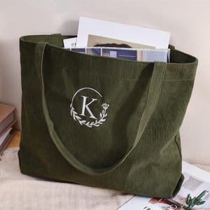 May include: Olive green corduroy tote bag with a white embroidered initial "K" inside a floral wreath. The bag is filled with books and papers. The tote has long handles for carrying.