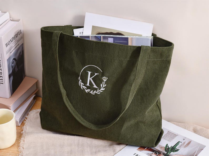 May include: Olive green corduroy tote bag with a white embroidered initial "K" inside a floral wreath. The bag is filled with books and papers. The tote has long handles for carrying.