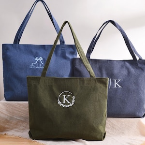 May include: Three corduroy tote bags in shades of blue and olive green. The bags have long handles and embroidered details, including a bow and the name "&Eacute;milie", a letter "K" inside a wreath, and the letter "K".