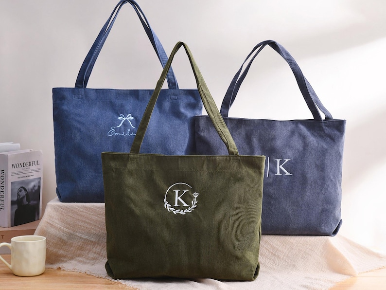May include: Three corduroy tote bags in shades of blue and olive green. The bags have long handles and embroidered details, including a bow and the name "&Eacute;milie", a letter "K" inside a wreath, and the letter "K".