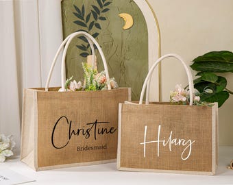 May include: Two brown burlap tote bags with white handles. The bag on the left has the name "Christine" and the word "Bridesmaid" printed in black. The bag on the right has the name "Hilary" printed in white.