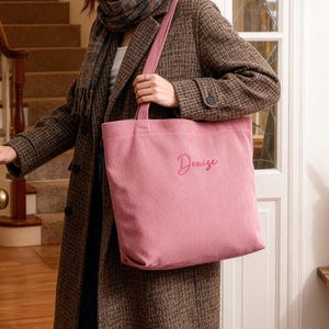 May include: A pink corduroy tote bag with the name "Denise" embroidered in a cursive font. The bag has a rectangular shape with a long strap. The bag is being held by a person wearing a brown coat.
