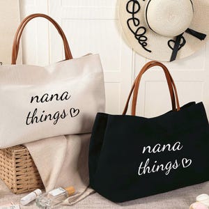 Può includere: Due borse tote con manici in pelle marrone. Una borsa è bianca con testo nero che dice "nana things" con un cuore. L'altra borsa è nera con testo bianco che dice "nana things" con un cuore.
