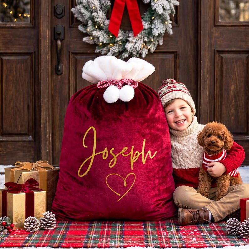 May include: A large, deep red velvet Christmas gift sack with a white faux fur trim and drawstring closure. The sack features the name Joseph and a heart icon in gold script. It is positioned next to gold gift boxes with red velvet ribbons.