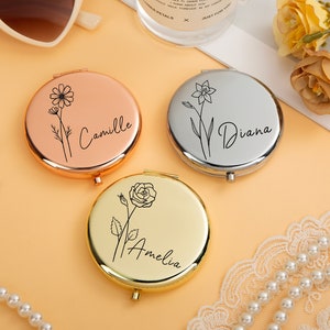 May include: Three round metal compact mirrors in rose gold, silver, and gold finishes. Each mirror features a black line art floral illustration and a name engraved in script: Camille with a daisy, Diana with a daffodil, and Amelia with a rose.