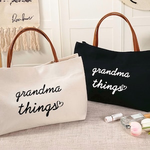 Può includere: Due borse tote in tela con manici in pelle marrone. Le borse sono bianche e nere con il testo "grandma things" stampato in nero.