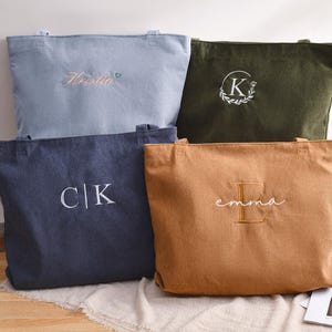 K&ouml;nnte beinhalten: Vier Cord-Tote-Bags in verschiedenen Farben: Hellblau, Olivgr&uuml;n, Marineblau und Beige. Jede Tasche hat gestickten Text oder Initialen in Wei&szlig;. Die Taschen werden auf einer neutralen Oberfl&auml;che pr&auml;sentiert.