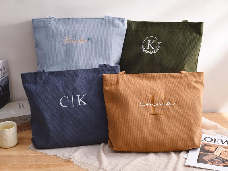 K&ouml;nnte beinhalten: Vier Cord-Tote-Bags in verschiedenen Farben: Hellblau, Olivgr&uuml;n, Marineblau und Beige. Jede Tasche hat gestickten Text oder Initialen in Wei&szlig;. Die Taschen werden auf einer neutralen Oberfl&auml;che pr&auml;sentiert.
