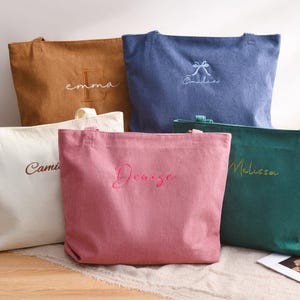 May include: Five corduroy tote bags in various colors: tan, blue, white, pink, and green. Each bag has a name embroidered on it in a contrasting color. The bags are rectangular with handles.