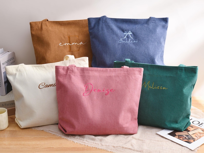 May include: Five corduroy tote bags in various colors: tan, blue, white, pink, and green. Each bag has a name embroidered on it in a contrasting color. The bags are rectangular with handles.
