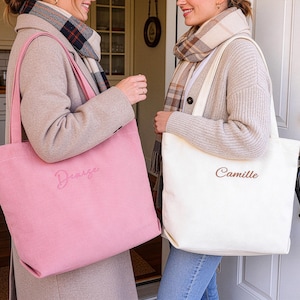 May include: Two tote bags, one pink and one off-white, are held by two people. The pink bag has the name "Denize" embroidered on it, while the off-white bag has "Camille". Both bags have long handles.