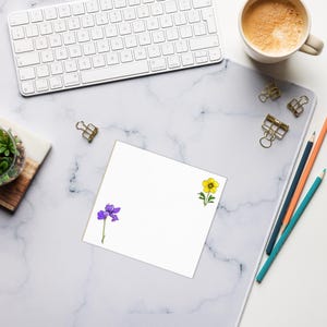 May include: A white keyboard, coffee cup, and stationery items are arranged on a marble-patterned desk mat. A blank white notepad features a purple iris and a yellow flower illustration. Gold binder clips and colored pencils are also present.
