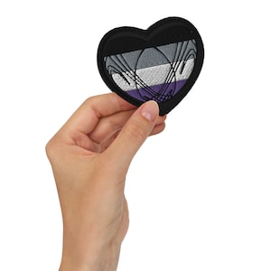 May include: A heart-shaped patch with a black border and an asexual pride flag design. The flag features horizontal stripes of black, grey, white, and purple. The patch is held in a hand.