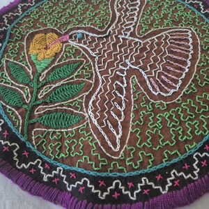 May include: A circular textile wall hanging featuring brown fabric with intricate white embroidered outlines of a bird and green embroidered floral motifs. The piece is bordered by a black band with pink cross-stitch details and a purple fringe.