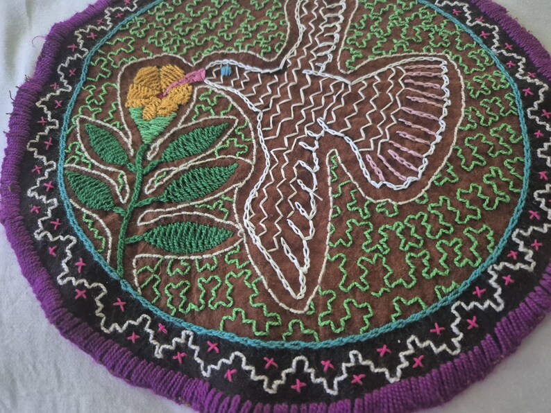 May include: A circular textile wall hanging featuring brown fabric with intricate white embroidered outlines of a bird and green embroidered floral motifs. The piece is bordered by a black band with pink cross-stitch details and a purple fringe.