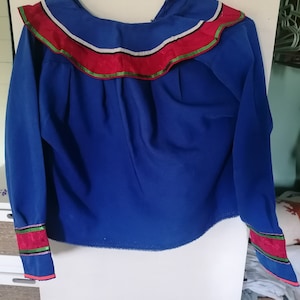 Könnte beinhalten: Ein blaues Langarmhemd mit einem Rüschenkragen und roten, weißen und grünen Verzierungen. Das Hemd hat einen dekorativen Besatz an den Manschetten.