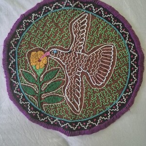 May include: A circular embroidered textile patch featuring a brown background with a green maze-like pattern. A white-outlined bird with a pink beak is shown flying towards a yellow flower with green leaves. The border is black with purple fringe.