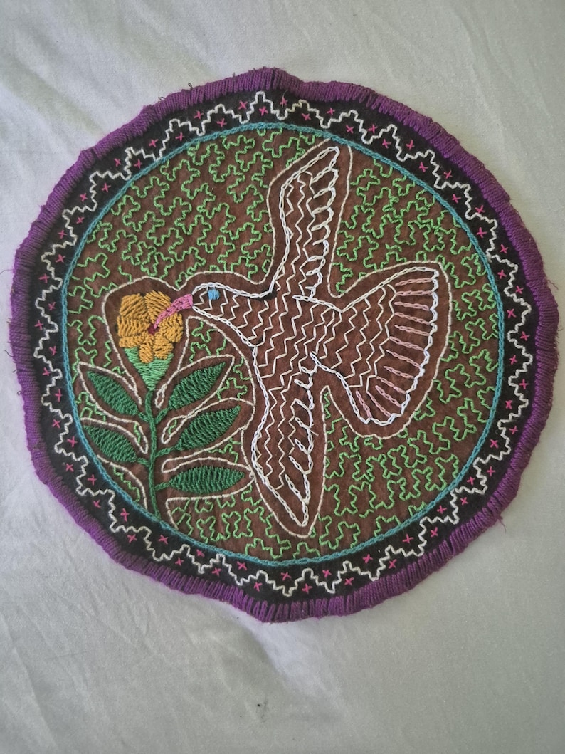 May include: A circular embroidered textile patch featuring a brown background with a green maze-like pattern. A white-outlined bird with a pink beak is shown flying towards a yellow flower with green leaves. The border is black with purple fringe.