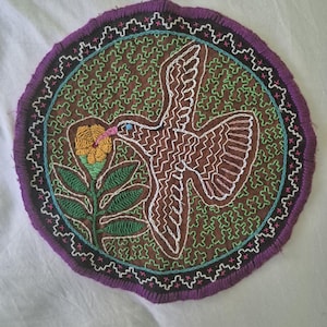 Op de afbeelding: Een ronde geborduurde textielpatch met een bruine achtergrond en een groen geometrisch patroon. Een vogel met witte omlijning is afgebeeld in vlucht naar een gele bloem met groene bladeren. De rand is paars met witte en roze stiksels.