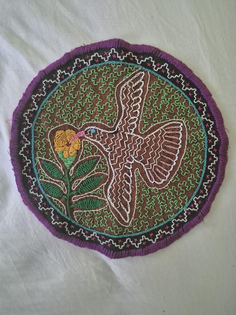 May include: A circular embroidered textile patch featuring a brown background with a green geometric pattern. A white-outlined bird is depicted in flight toward a yellow flower with green leaves. The border is purple with white and pink stitching.