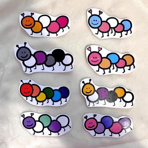May include: Eight colorful caterpillar stickers with different pride flag colors. Each sticker has a smiling face and two antennae.