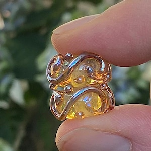 May include: A small, cylindrical glass bead featuring a translucent amber-colored core encased in a decorative, polished silver-toned metal wire overlay that forms swirling, interconnected patterns across the surface of the bead.