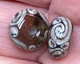 May include: Two glass beads with swirling patterns in shades of brown, white, and gray. One bead is oval-shaped, and the other is a smaller, rounded shape. The beads have a glossy finish and are held in a hand.