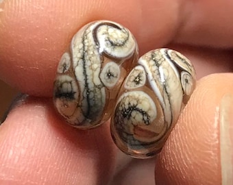 May include: Two glass beads with a swirling pattern of white, brown, and black. The beads are translucent and have a glossy finish.