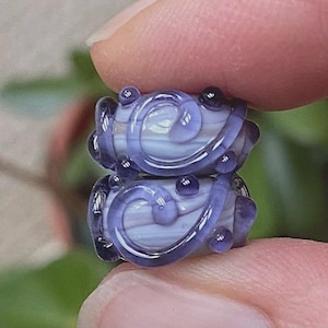 May include: Two cylindrical glass beads featuring a light lavender and white striped base. Each bead is decorated with a raised, dark purple glass swirl pattern that wraps around the surface, accented by small, round dark purple glass dots.