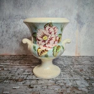 May include: Cream ceramic vase featuring a large pink and white floral design. The vase has a flared top, two small handles, and a pedestal base. The background is a muted blue-grey.