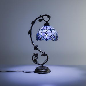 May include: A decorative table lamp with a stained glass shade in shades of blue and white. The lamp has a dark metal base and a curved stem with leaf accents. The shade has a floral pattern and is illuminated.
