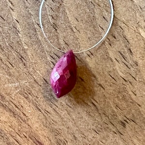 May include: A faceted, teardrop-shaped ruby gemstone pendant hangs from a thin silver wire. The ruby is a deep, rich red color with multiple facets that catch the light. The background is a textured, wooden surface.