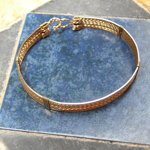 K&ouml;nnte beinhalten: Ein goldfarbenes Drahtarmband mit einem gewebten Muster. Das Armband hat einen einfachen Verschluss.