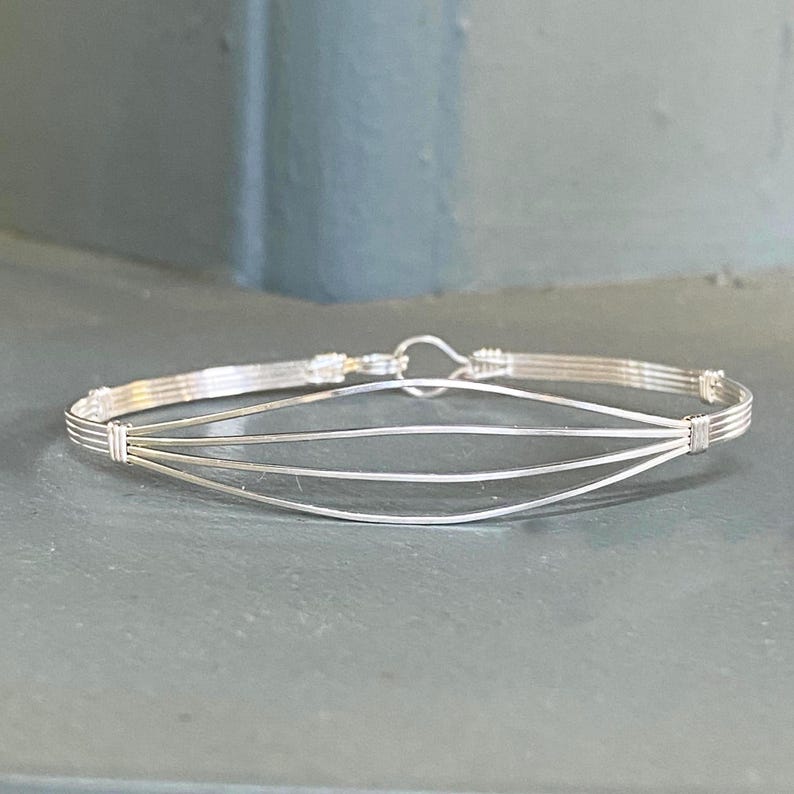 May include: A silver wire bracelet with a simple, elegant design. The bracelet is made of multiple strands of wire that are woven together to create a delicate and intricate pattern.