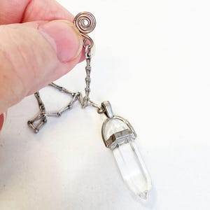 May include: A clear, faceted, hexagonal crystal point pendant suspended from a silver-toned metal chain. The chain features a decorative spiral-shaped metal clasp at the top, designed for secure attachment. The crystal is held by a silver-toned metal cap.