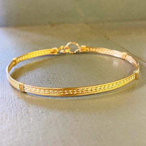 May include: A gold-toned bangle bracelet with a braided design. The bracelet is made of thin wire and has a simple clasp.