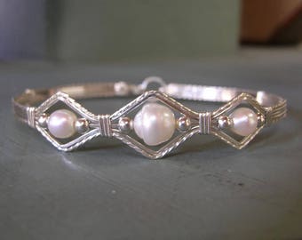 Könnte beinhalten: Ein silbernes, drahtgewickeltes Armband mit drei weißen Perlen, die in rautenförmigen Drahtdesigns eingefasst sind.