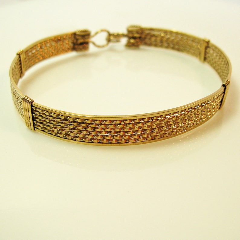 K&ouml;nnte beinhalten: Ein goldfarbenes Metallarmband mit gewebtem Design. Das Armband hat einen einfachen Verschluss.
