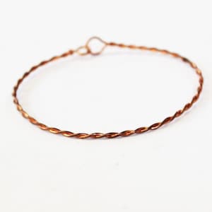 May include: A delicate copper-colored bracelet with a twisted wire design. The bracelet is circular, with a small loop closure. The thin wire is tightly wound, creating a textured appearance. The jewelry is set against a white background.