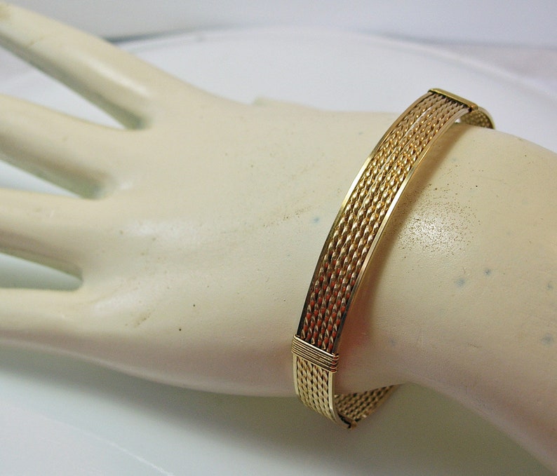 K&ouml;nnte beinhalten: Ein geflochtenes Drahtarmband in Goldton. Das Armband besteht aus mehreren Dr&auml;hten, die miteinander verflochten sind, um ein strukturiertes Muster zu erzeugen.